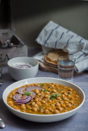 Channa masala or chole masala displayed in a white bowl with raita, roti and a glass of water.